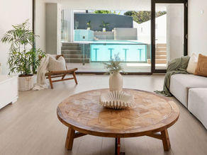 Modern living room with a wooden table, white vase, and sofa. Glass doors lead to a pool. Neutral tones and potted plant enhance calm. 