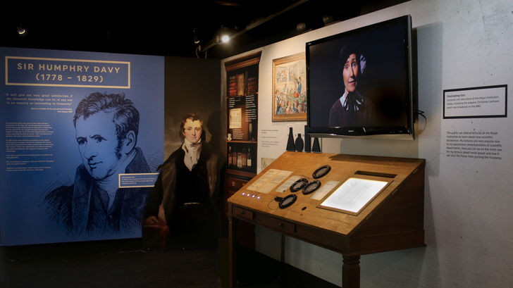 Exhibition space with graphic panels of text and imagery including an illustration of Sir Humphrey Davy. An interactive desk with bottles and magnifying glass. A screen with a lady shown on it hangs above the table.