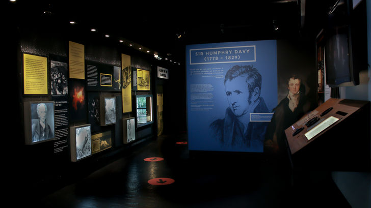 Exhibition space with orange arrows marking the journey. Graphic panels with images and text including large image of Sir Humphrey Davy. A table on the right featuring interactives and screen above.