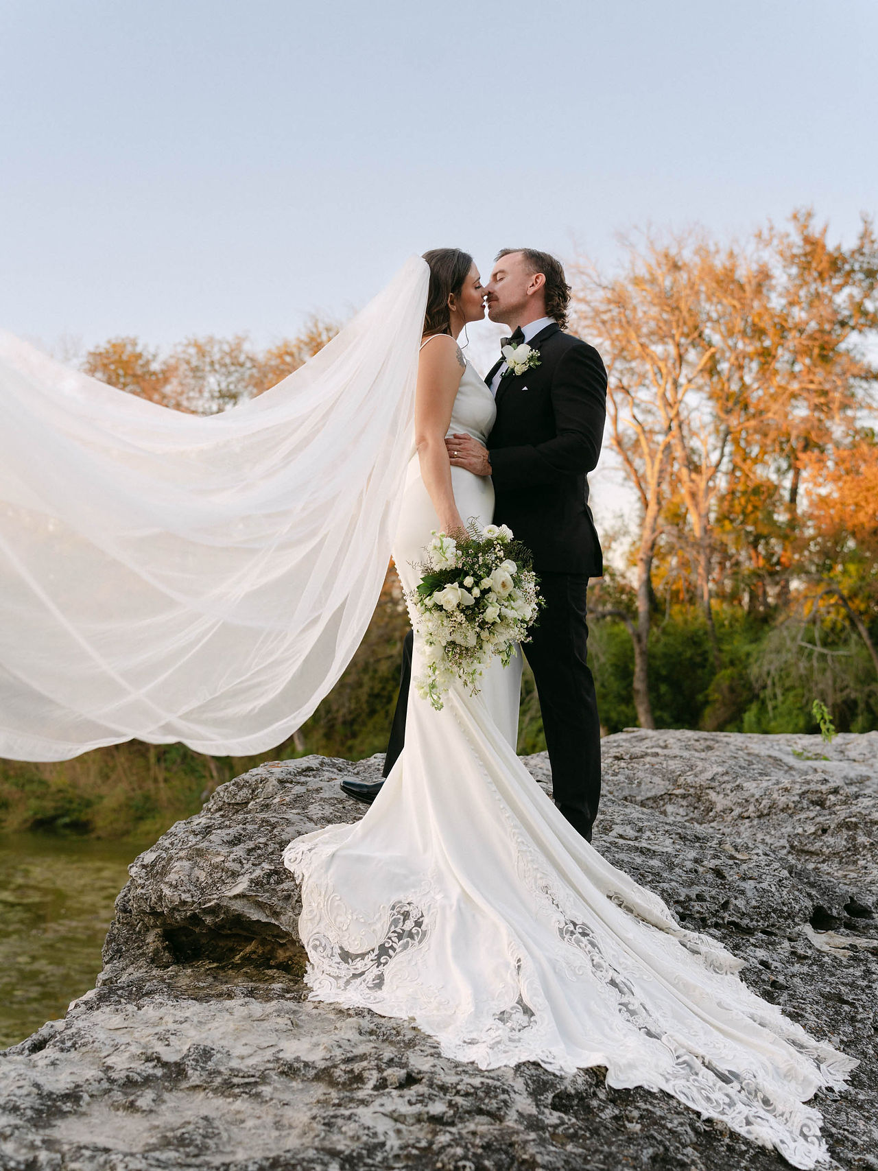 Bride and groom share a kiss as her veil flows in the wind, elegant Texas wedding portrait captured at sunset in soft, natural light.