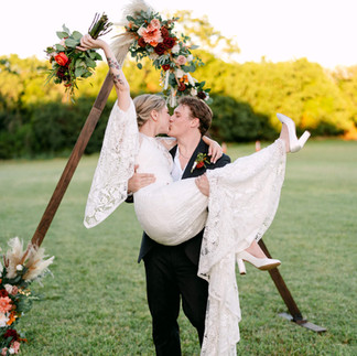 Groom lifts bride in celebration beneath a floral arch, joyful outdoor Texas wedding moment filled with laughter and color.