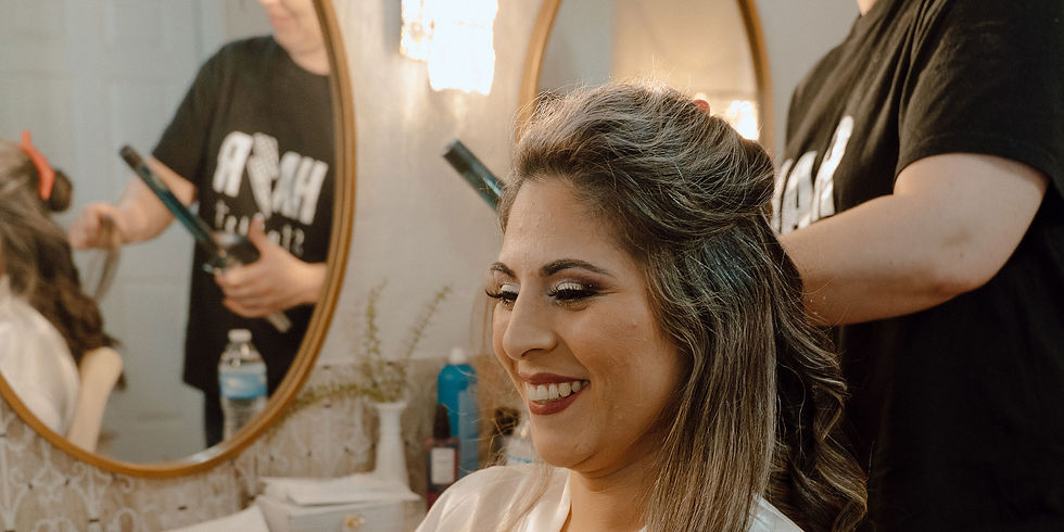 Bride smiling while getting her hair styled with a curling iron in a light and airy bridal suite. Mirror reflects stylist. Bright, cozy setting at a wedding venue near Rosenberg TX