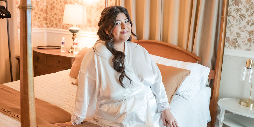 Woman in a white robe sits on a bed in a cozy room with floral wallpaper and soft lighting, smiling gently.