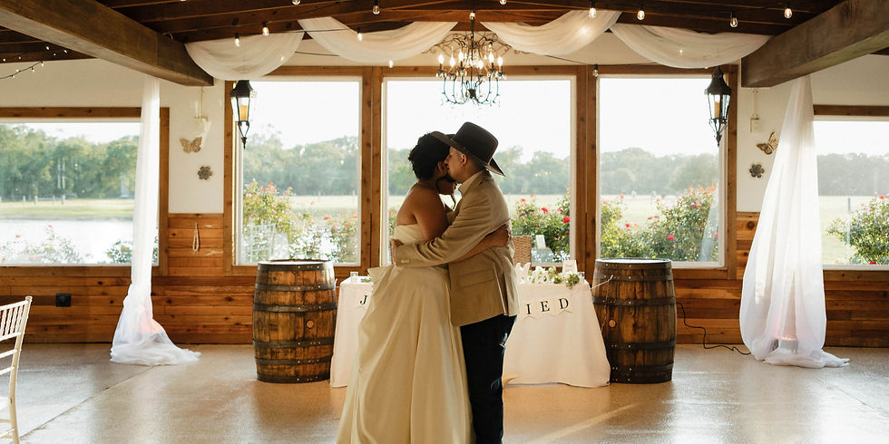 A newly-wed couple kisses in a rustic reception hall with wooden barrels, white drapes, and a "Just Married" sign. Sunlight streams through large windows at a wedding venue near Cypress TX
