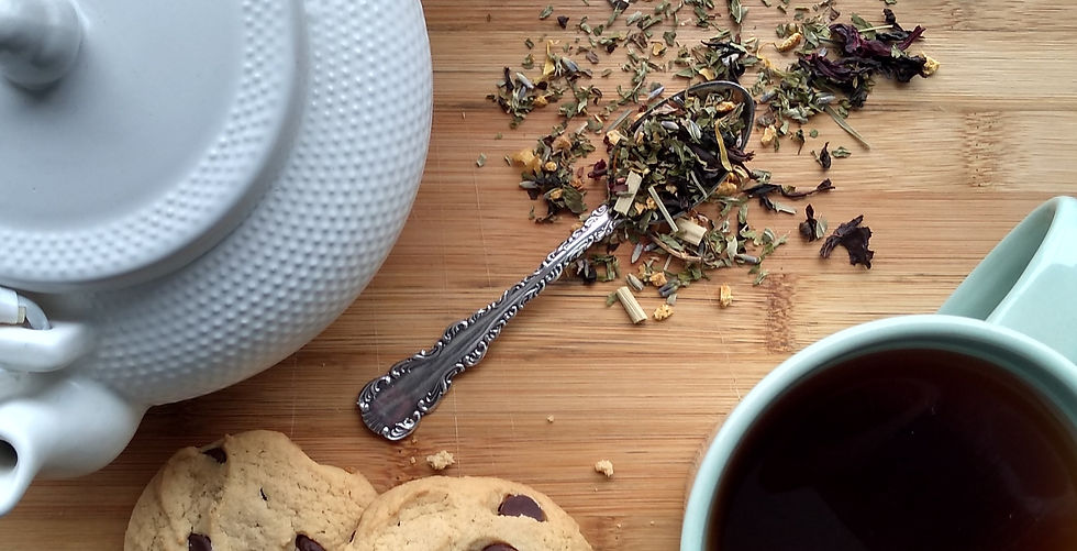 A teaspoon full of loose-leaf tea lays on a wooden cutting board next to a white teapot, a mug full of tea and 2 chocolate chip cookies