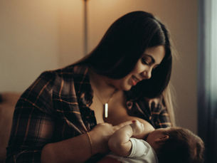 A dark haired woman sits in a warmly lit room breastfeeding an older infant