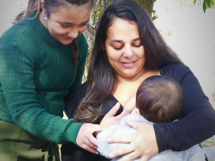 A woman in green leans over the shoulder of a woman in black who is nursing a baby. The woman in green is assisting with the breastfeeding.