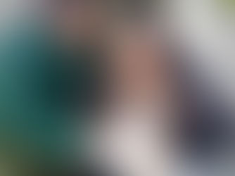 A woman in green leans over the shoulder of a woman in black who is nursing a baby. The woman in green is assisting with the breastfeeding.