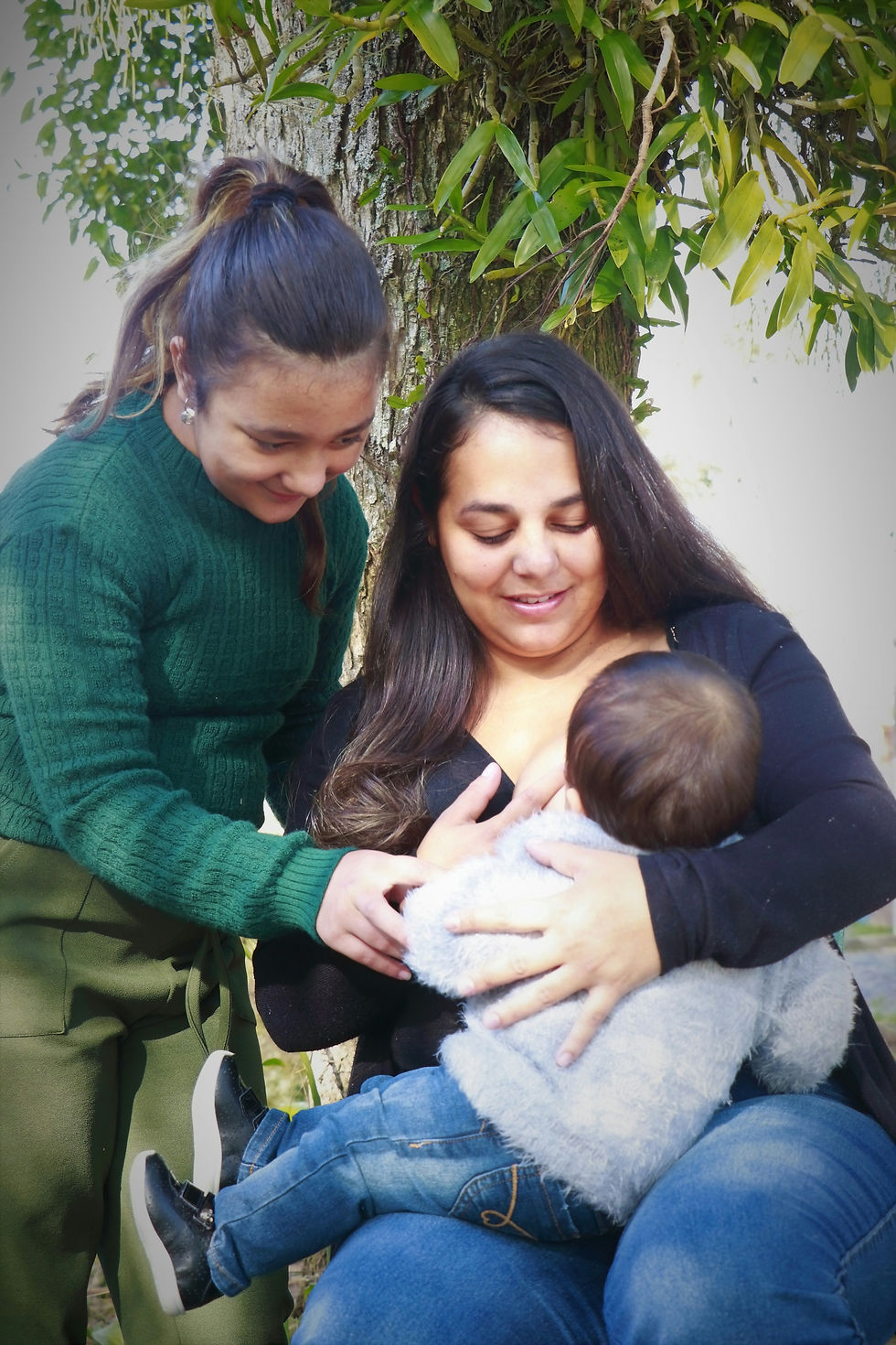 A woman in green leans over the shoulder of a woman in black who is nursing a baby. The woman in green is assisting with the breastfeeding.