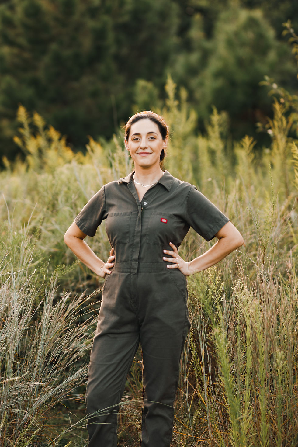 A woman in a dark green jumpsuit stsands in a field of tall grass backlit by sunshine.
