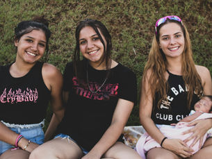 Three women sitting against a grassy background, the rightmost woman holds a small infant.