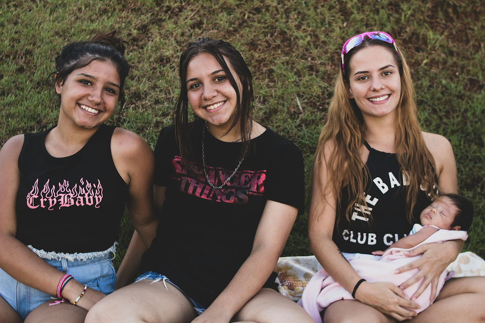 Three women sitting against a grassy background, the rightmost woman holds a small infant.