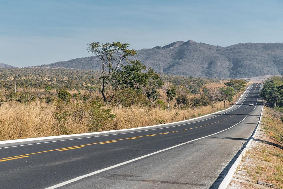 Estrada asfaltada vazia em paisagem seca montanhosa, com um carro ao longe sob céu azul claro.