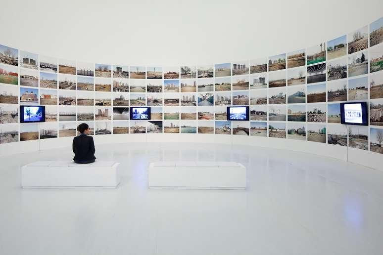 Pessoa sentada em banco branco numa galeria minimalista, diante de parede curva com fotos e telas, em clima silencioso.