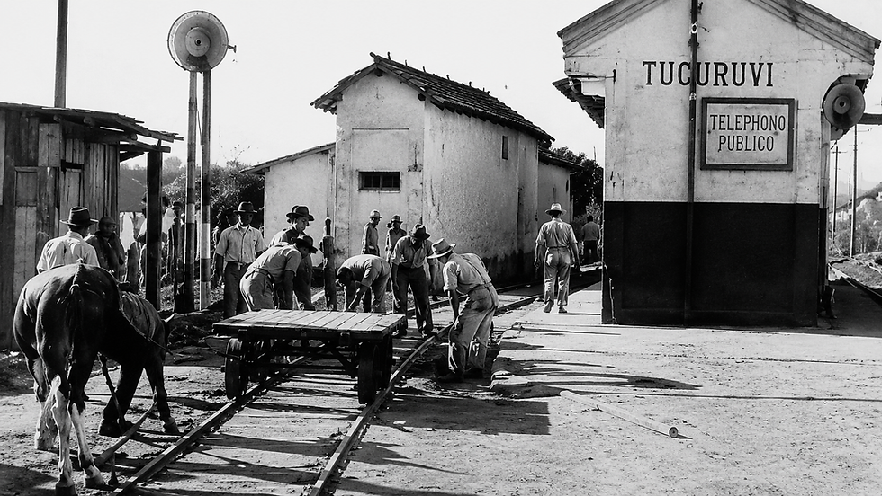 Homens e cavalo ao lado de trilhos, em rua de terra, diante de prédio com placas TUCURUVI e TELEPHONO PUBLICO, cena antiga.