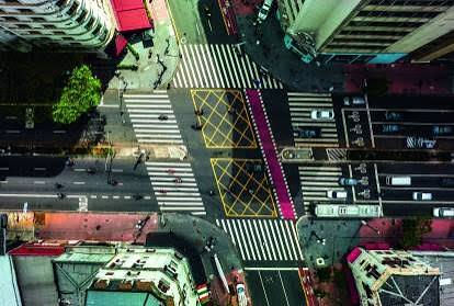 Vista aérea de cruzamento urbano com faixas de pedestres, carros e pessoas; marcações amarelas e faixa rosa destacam o asfalto.
