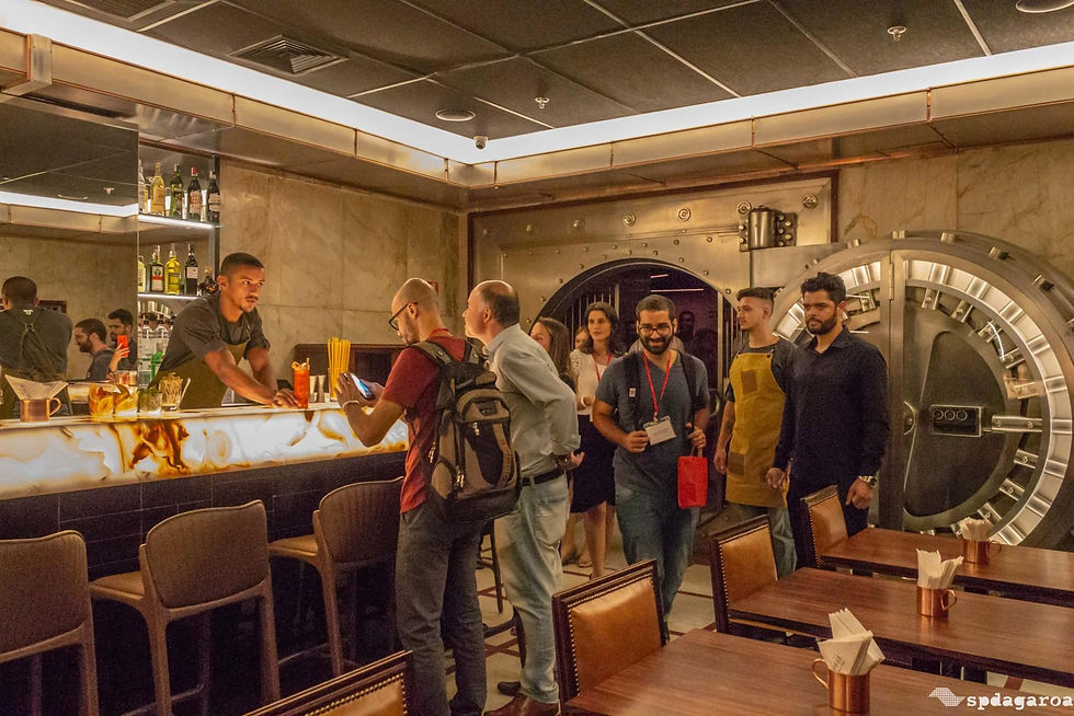 Grupo em bar sofisticado, com balcão iluminado, coquetéis e cofre metálico ao fundo, em clima animado.