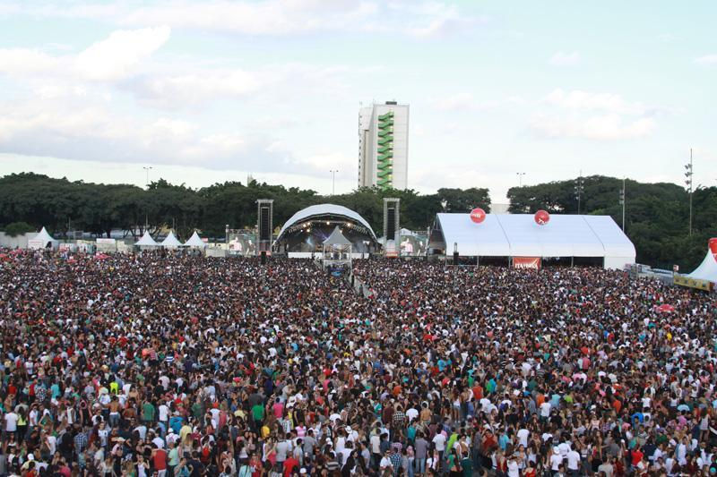 Multidão enorme em festival ao ar livre, diante de palco e tendas brancas, sob céu claro e clima animado.