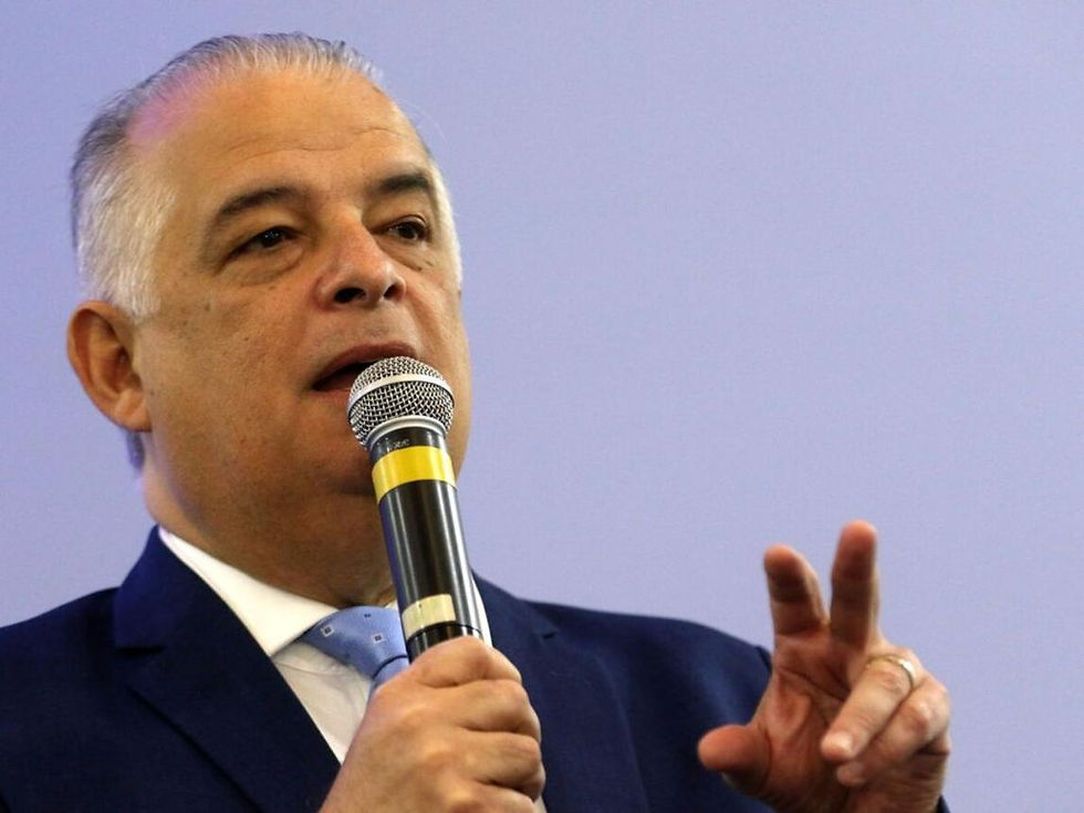 Homem de terno fala ao microfone e gesticula com a mão, diante de fundo azul claro.