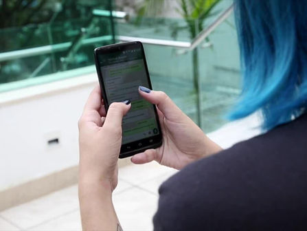 Pessoa de cabelo azul usa celular e envia mensagens em app de chat, em ambiente interno com corrimão de vidro.