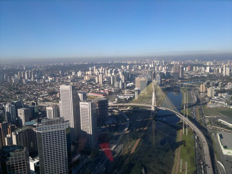 Vista aérea de grande cidade com ponte estaiada sobre rio, prédios altos e céu azul limpo.
