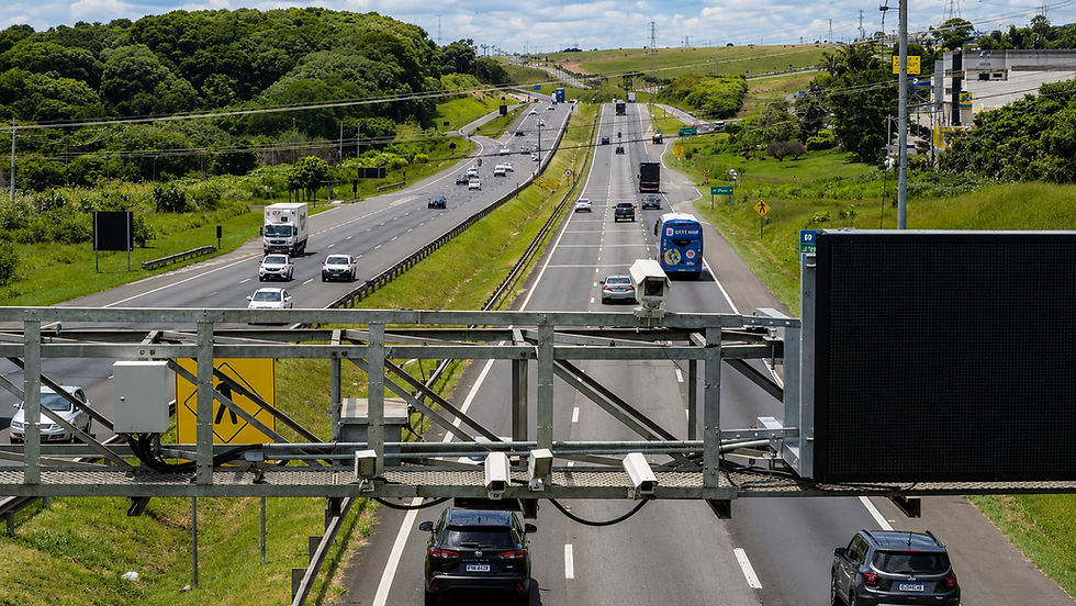 Rodovia dupla com carros e ônibus, cercada por morros verdes; painel eletrônico vazio em primeiro plano.
