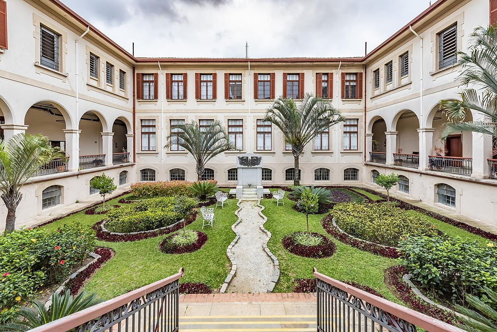 Pátio interno de prédio histórico com jardim bem cuidado, palmeiras, bancos brancos e fachada creme com arcos e janelas.