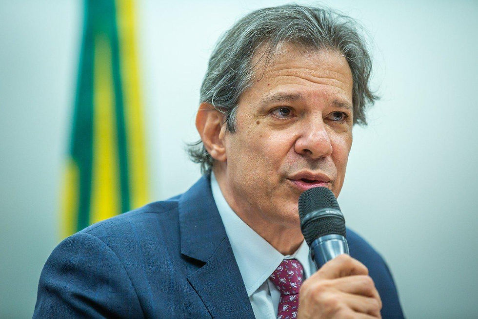 Homem de terno fala ao microfone, com bandeira do Brasil desfocada ao fundo.