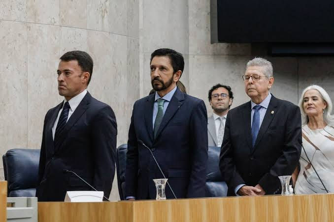 Quatro autoridades de terno em plenário, de pé e sérias, diante de mesa de madeira e cadeiras azuis.