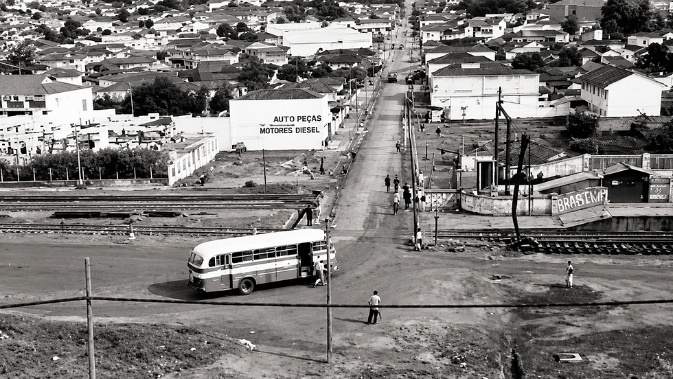Ônibus parado em cruzamento junto a trilhos, pessoas andando num bairro urbano; placa AUTO PEÇAS MOTORES DIESEL ao fundo.