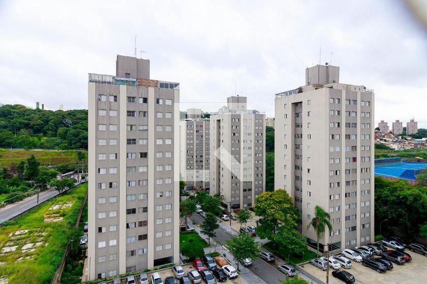 Condomínios altos cinza em área urbana, com estacionamento e árvores sob céu nublado; ícone de play no centro.