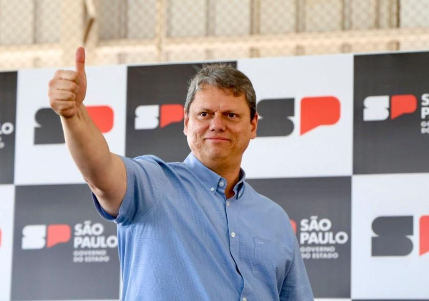 Homem de camisa azul faz sinal de positivo diante de painel com logos de São Paulo; expressão confiante.