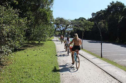 Três ciclistas pedalando por uma trilha em parque ensolarado, com árvores, gramado e estrada vazia ao lado.