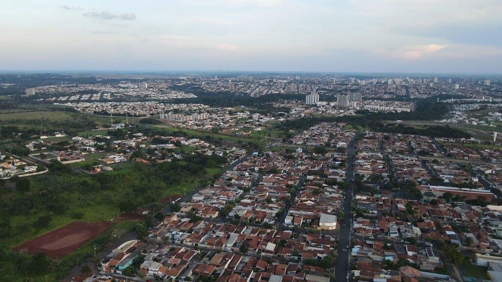 Vista aérea de bairro residencial com casas de telhados vermelhos, áreas verdes e skyline urbano sob céu nublado ao entardecer.