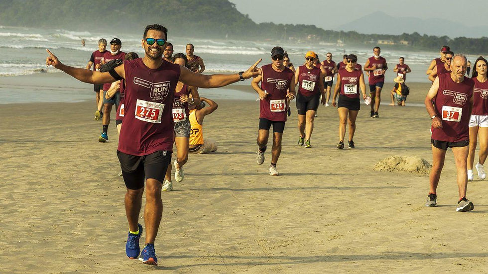 Corredores de camiseta vinho correm na praia; homem à frente sorri de braços abertos, com mar e montanhas ao fundo.