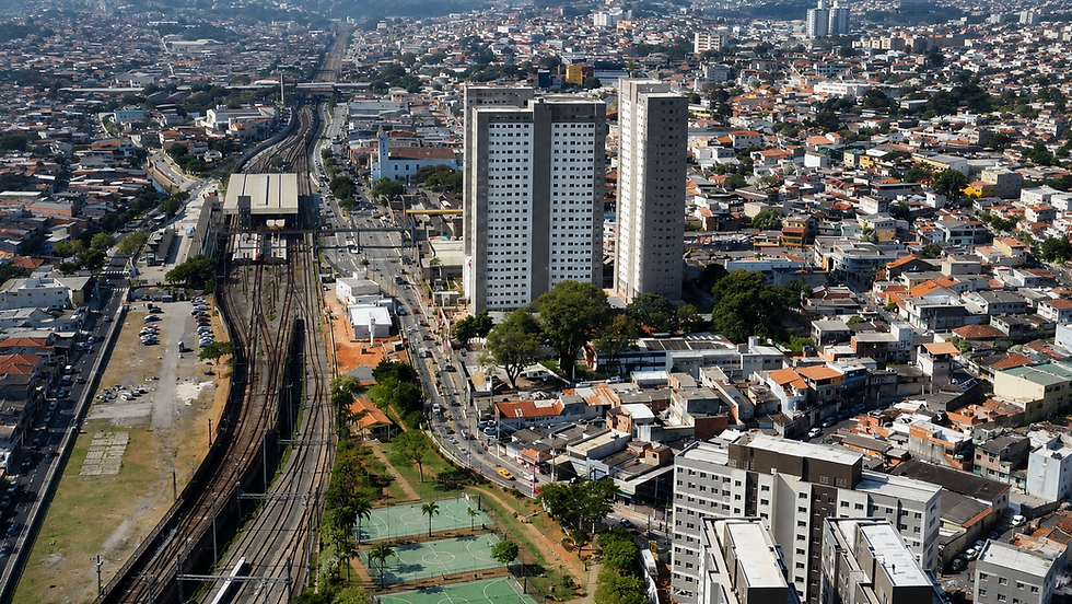 Vista aérea de cidade com trilhos de trem, torres altas, casas densas e quadras esportivas sob céu claro.