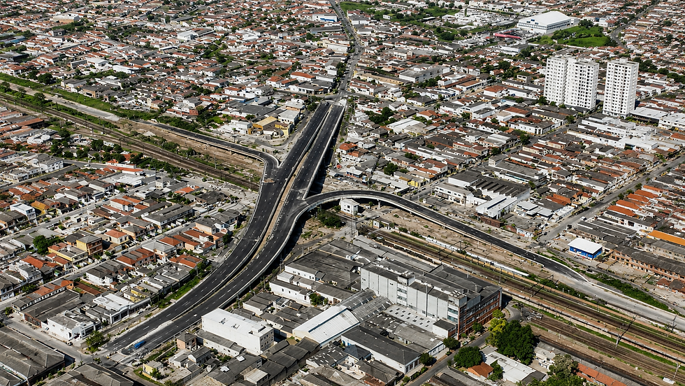Vista aérea de cidade com viaduto e cruzamento de avenidas, casas de telhas vermelhas e linhas férreas ao fundo.