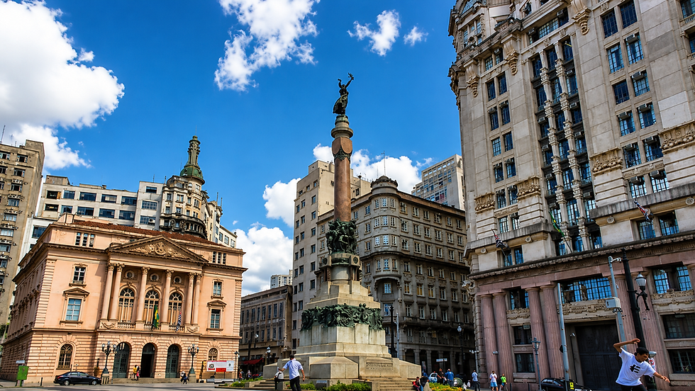 Praça urbana com monumento central, prédios históricos ao redor e céu azul com nuvens; poucas pessoas caminhando.