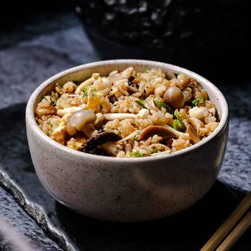 Tigela de arroz frito com cogumelos, em louça bege sobre pedra escura, com hashis ao lado.