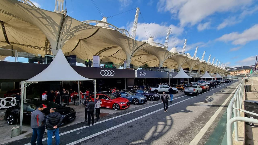 Box de pit lane com carros esportivos Audi e Ford, pessoas sob tendas brancas e céu azul ensolarado.