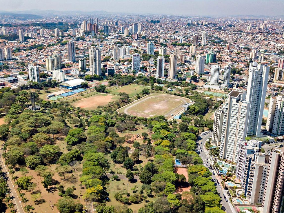 Vista aérea de cidade com arranha-céus, parque arborizado e pista de atletismo; dia claro e agitado.