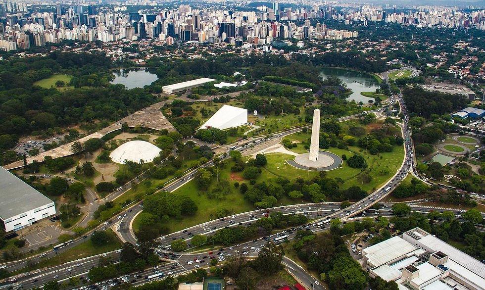 Vista aérea de parque urbano com obelisco, lagos, vias movimentadas e prédios ao fundo sob céu claro.