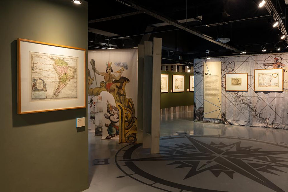Sala de museu com mapas antigos emoldurados, painéis ilustrados e chão com rosa dos ventos, sob luz baixa.