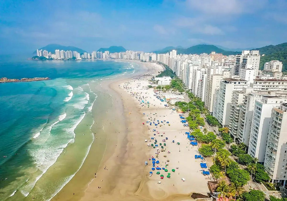 Vista aérea de praia urbana com mar azul, ondas, guarda-sóis coloridos e arranha-céus à beira-mar sob céu claro.