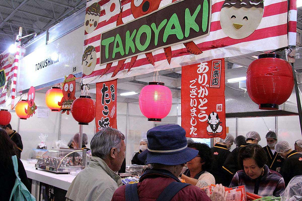 Barraca de takoyaki com lanternas vermelhas e cartazes japoneses; clientes compram em ambiente de feira movimentada.