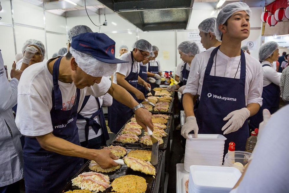 Cozinheiros de touca e avental azul preparam panquecas na chapa em barraca movimentada, com pilhas de pratos e clima de feira.