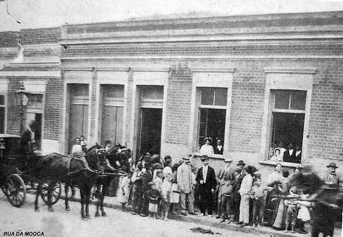 Rua da Mooca em foto antiga: multidão, carroça puxada por cavalos e pessoas diante de prédio, clima histórico.