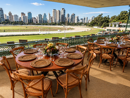 Vista deslumbrante do Jockey Club, destacando mesas elegantemente preparadas para eventos, com o hipódromo ao fundo e a impressionante silhueta urbana de São Paulo.