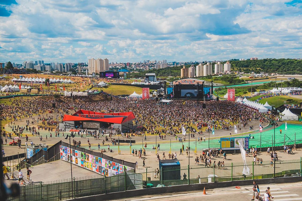 Multidão em festival ao ar livre com palco, tendas e placa Bud Station; céu nublado e skyline urbano ao fundo.