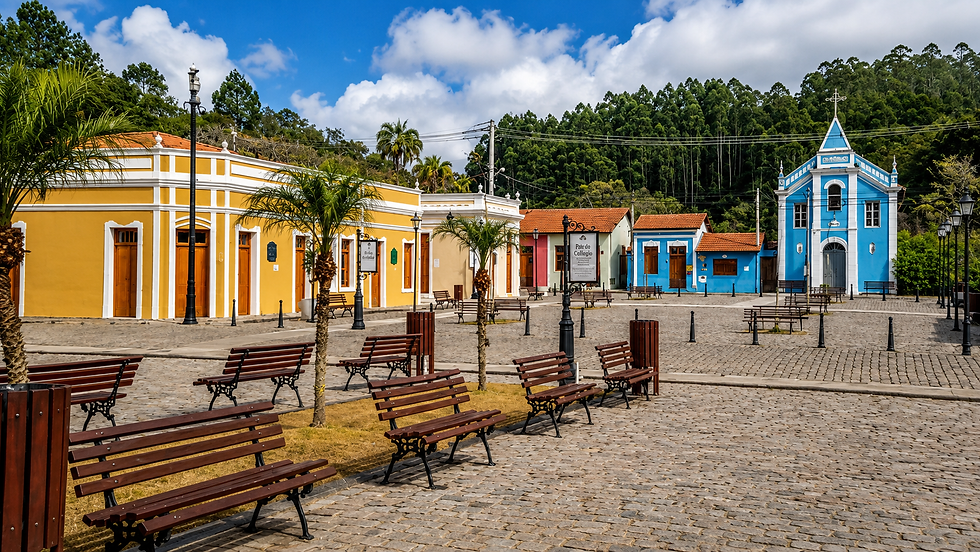 Praça de paralelepípedos com bancos, casas coloridas e uma igreja azul ao fundo, sob céu ensolarado.
